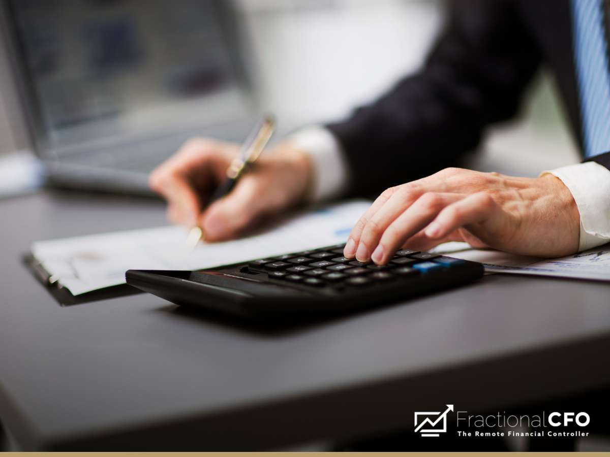 A Fractional CFO at work, using a calculator and writing notes, with a laptop in the background.